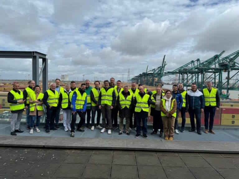 Group of people wearing safety vests at industrial port with cargo cranes and shipping containers in the background.