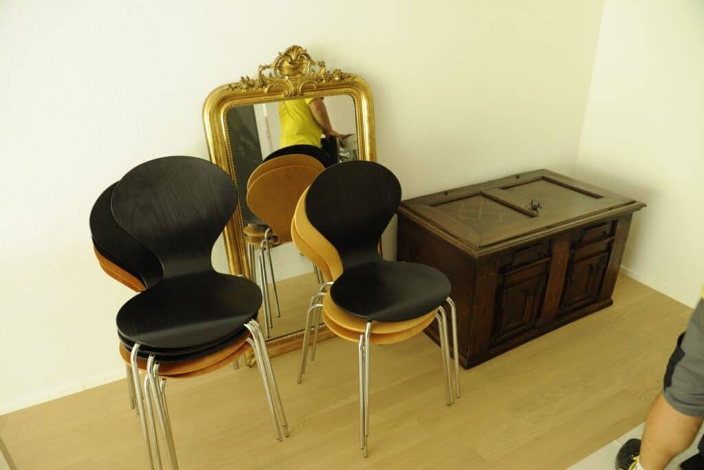 Stacked modern chairs with black, beige, and orange seats reflecting in a gold-framed mirror. A wooden cabinet is beside the chairs.