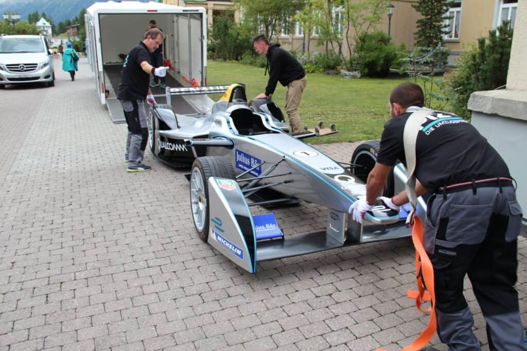High-performance race car being loaded on transport trailer by team members at a garage or paddock area.
